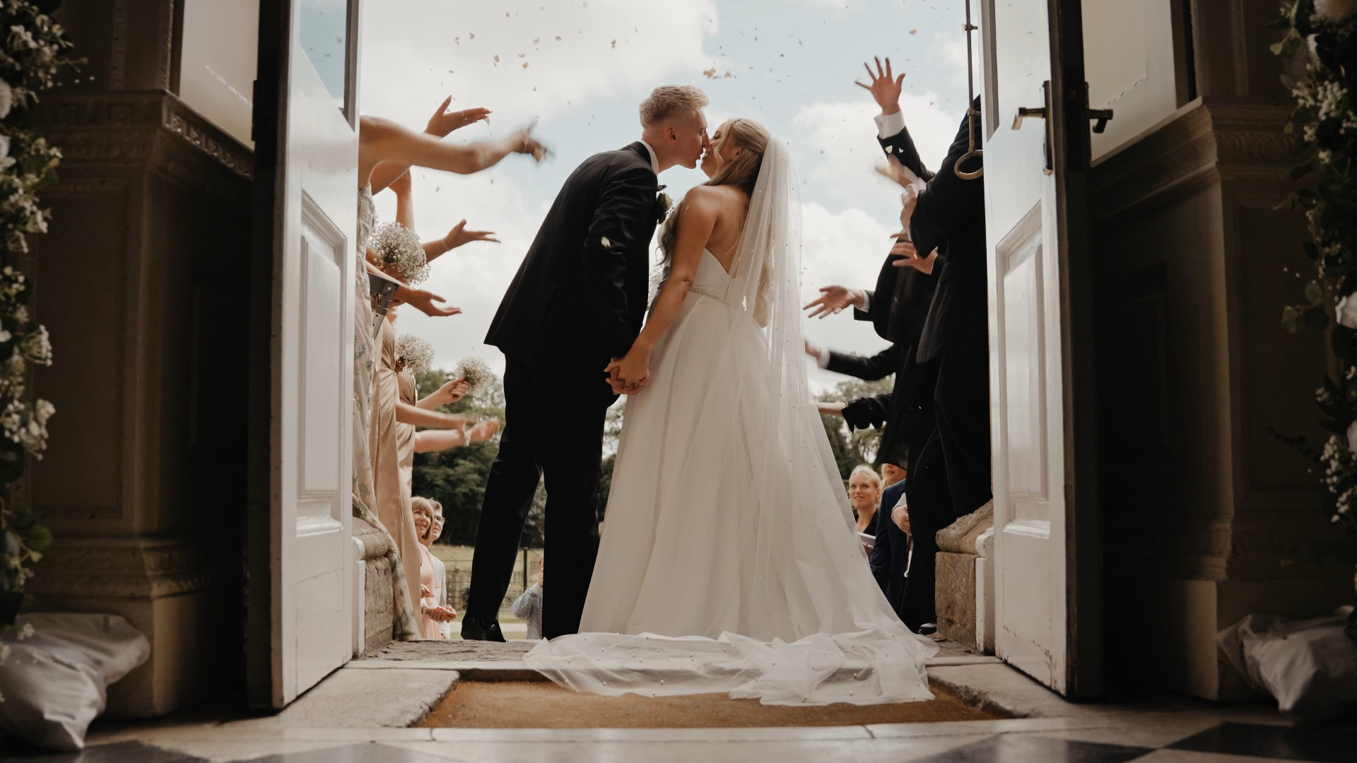 The bride and groom share a joyful kiss as guests cheer and toss flower petals around them, a vibrant and celebratory scene captured by the Best Wedding Videographer in the South West.