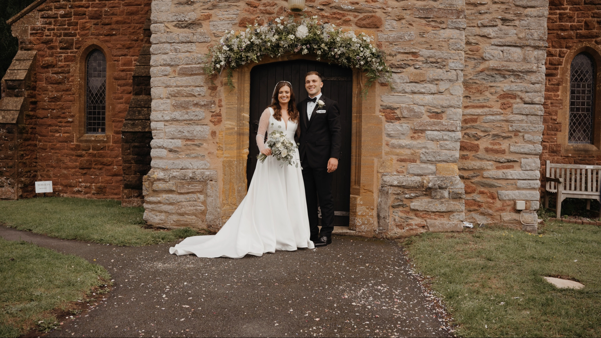 The bride and groom smile warmly at the camera while posing outside the wedding venue, capturing a joyful and elegant moment filmed by the Best Wedding Videographer in the South West.