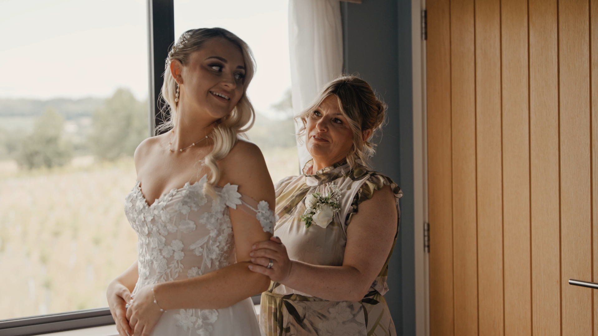 The bride looks back and smiles warmly at her bridesmaid who is buttoning up her dress, a tender and joyful moment captured by the Best Wedding Videographer in the South West.