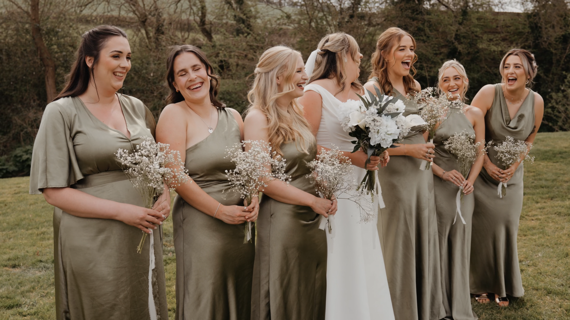 Bridal party in moss green dresses laughing and smiling with the bride during a wedding at Priston Mill.

