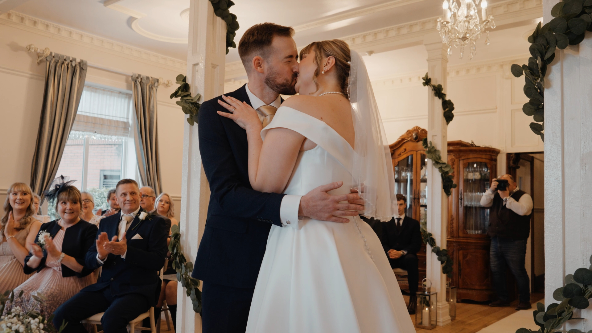 A bride and groom share a kiss at their indoor Berwick Lodge wedding ceremony. The bride wears a white off-shoulder gown and veil; the groom is in a dark suit. Guests smile and applaud in the elegantly decorated room.