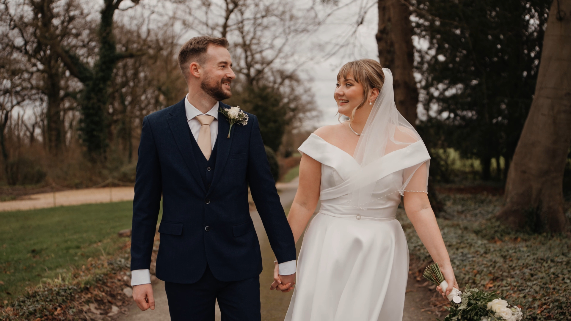 A bride in a white dress and veil and a groom in a navy suit hold hands and smile at each other while walking outdoors on a tree-lined path at Berwick Lodge.