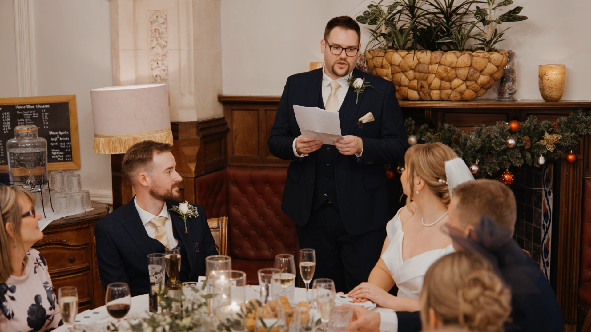A man in a suit stands and reads from a paper, giving a speech at an intimate wedding at Berwick Lodge wedding reception. Seated guests in formal attire, including the bride and groom, listen attentively at a decorated table with drinks and candles.