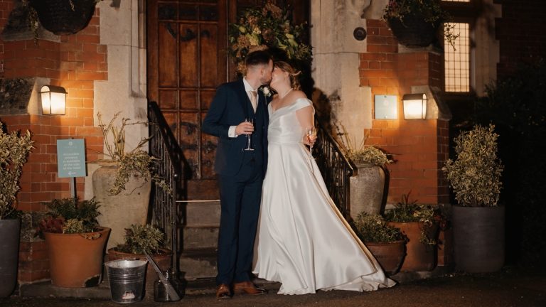 A bride and groom, dressed in a white gown and dark suit, share a kiss while holding champagne glasses in front of a brick building at night—an intimate wedding at Berwick Lodge.