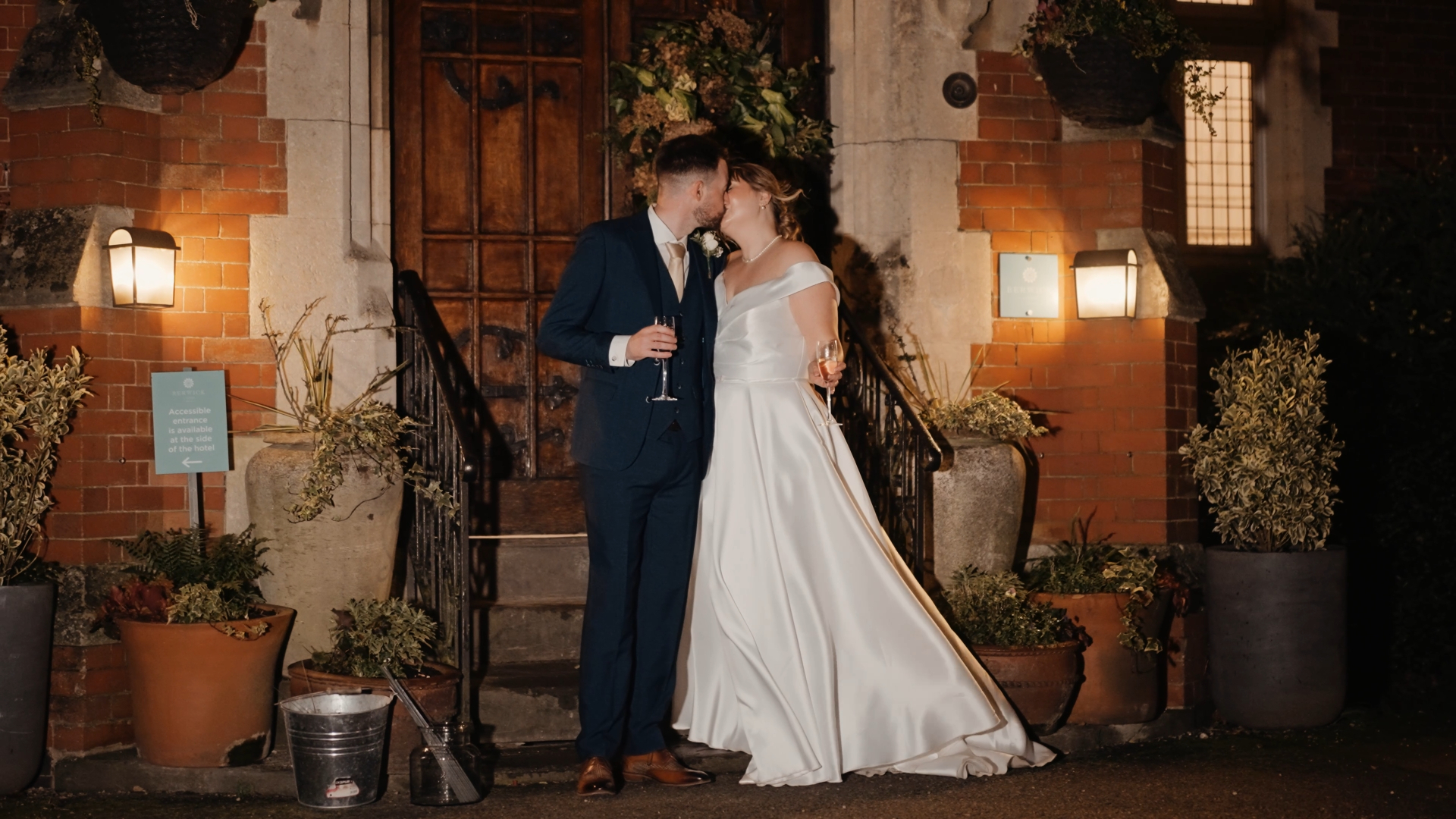 A bride and groom, dressed in a white gown and dark suit, share a kiss while holding champagne glasses in front of a brick building at night—an intimate wedding at Berwick Lodge.