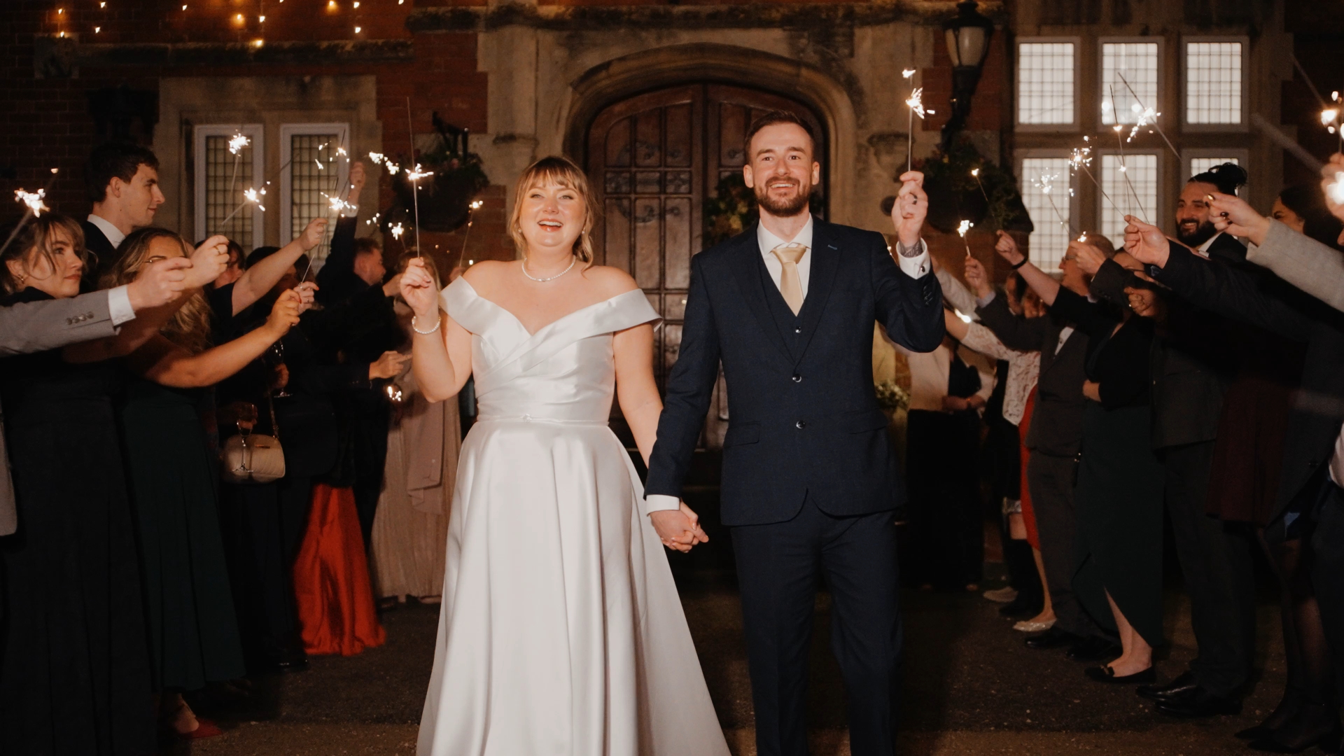 A newlywed couple, the bride in a white gown and the groom in a dark suit, hold hands and sparklers while smiling during their intimate wedding at Berwick Lodge, surrounded by guests with sparklers outside a building at night.