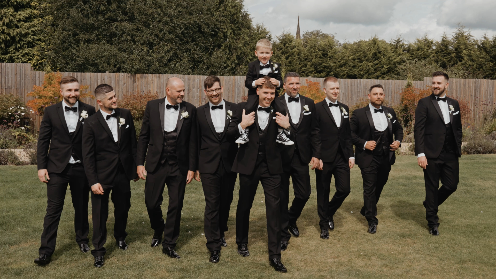 Groom walking in garden with groomsmen while his son enjoys a piggyback ride, a joyful moment captured in a wedding video package.