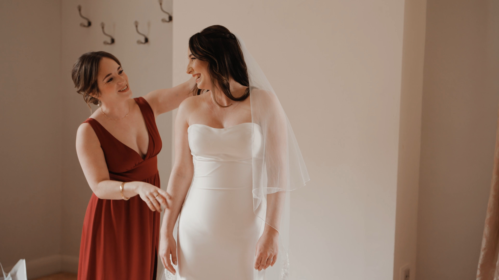 Bride smiling back at bridesmaid while having her dress buttoned, a candid moment captured in a wedding video package.