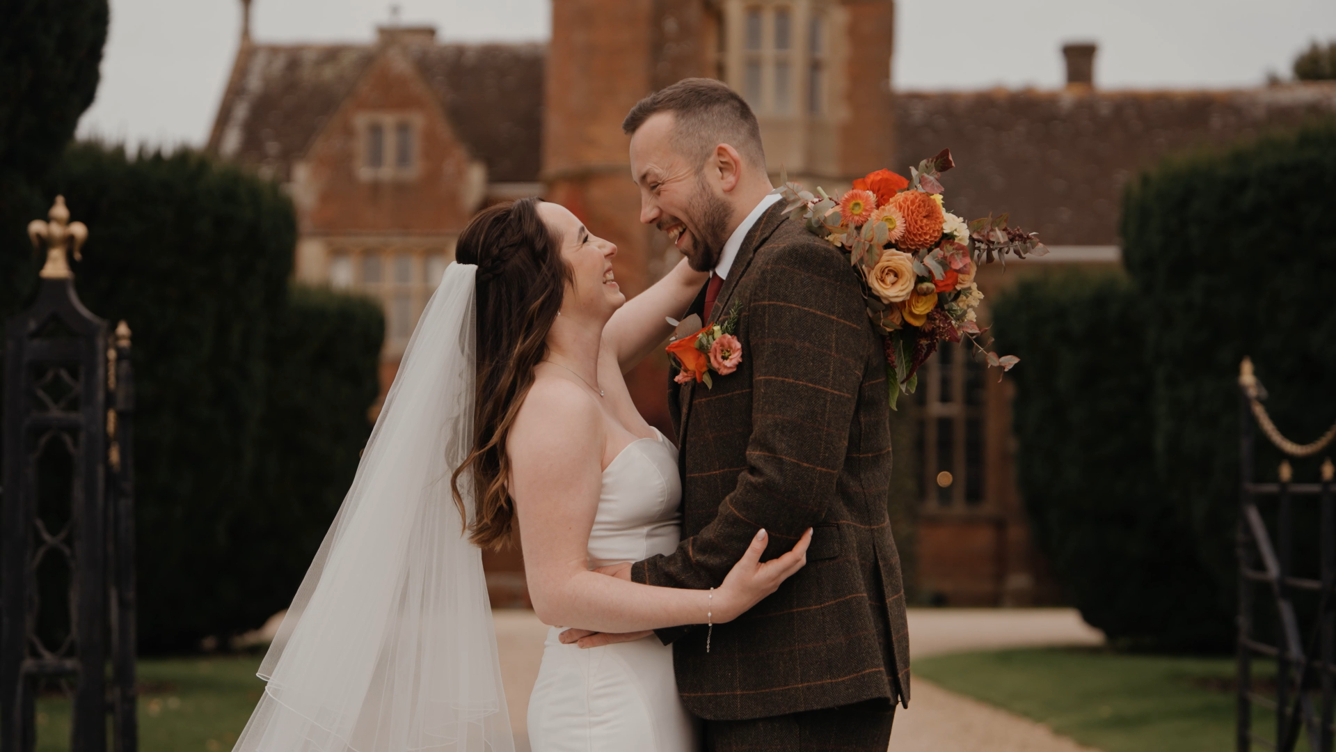 Bride wraps her hands around groom’s neck, holding a bouquet of burnt orange and red roses, perfect for autumn wedding styling tips.