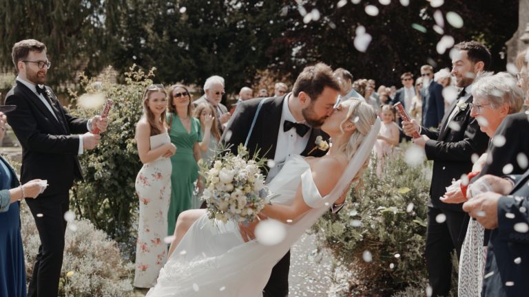 Groom dips bride for a romantic kiss as guests toss flower petals in a joyful celebration—captured like a cinematic wedding film