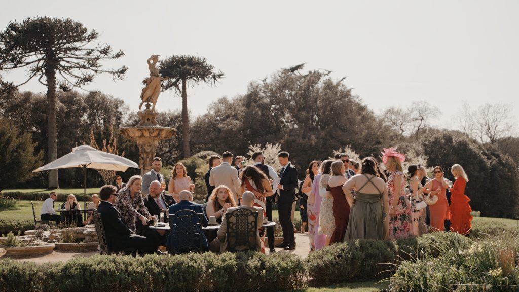 Wedding guests in vibrant, colorful attire mingle and chat outdoors at Clevedon Hall, surrounded by stunning landscape scenery under perfect weather during an intimate wedding celebration.