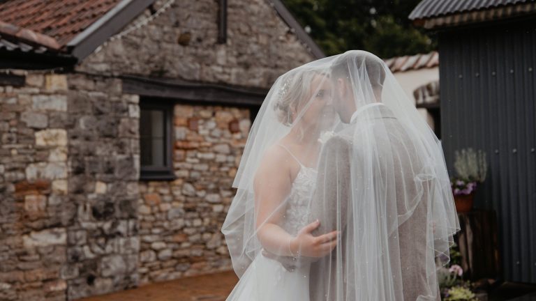 Bride and groom share a tender moment under the veil with the rustic stone buildings of Aldwick Estate Somerset behind them. A perfect rustic wedding venue setting.