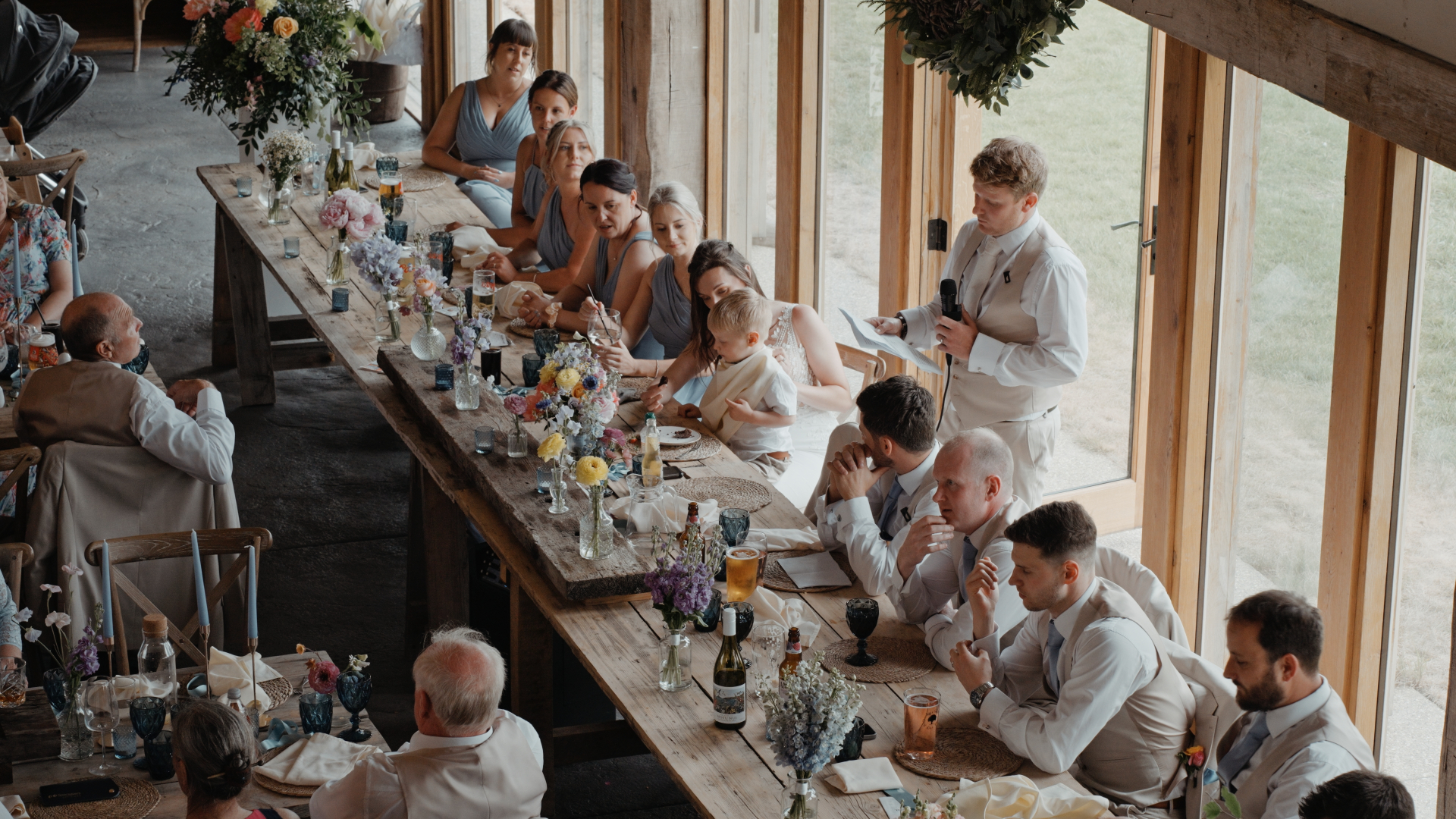 Guests seated at long rustic tables decorated with wildflowers and vintage glassware during wedding speeches inside Old Oak Farm.