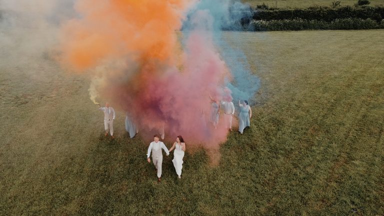 Bride and groom holding hands amid vibrant pink, orange, yellow, and purple smoke as wedding guests light smoke bombs, captured by a second videographer