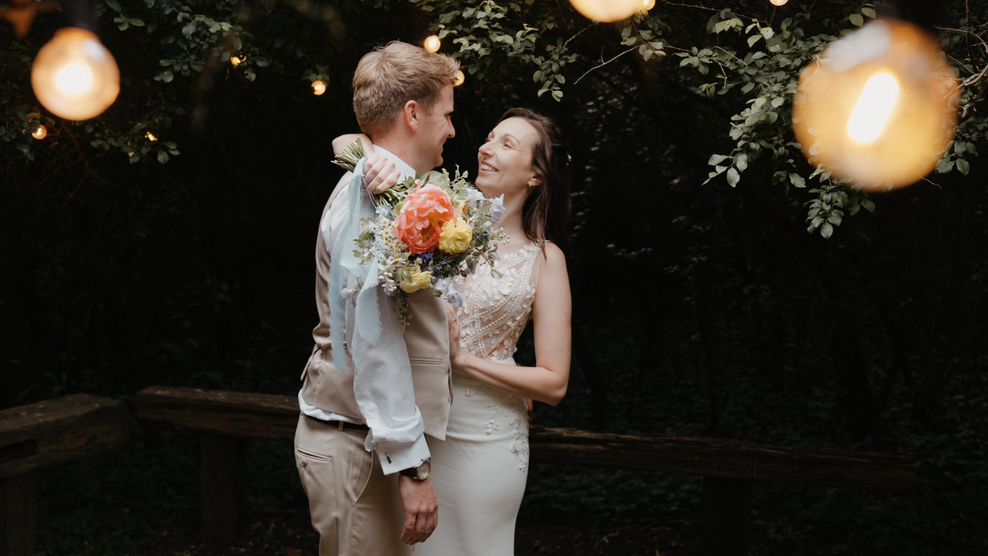 Bride hugs groom while holding a vibrant wildflower bouquet under warm fairy lights during a romantic moment at a Somerset wedding.