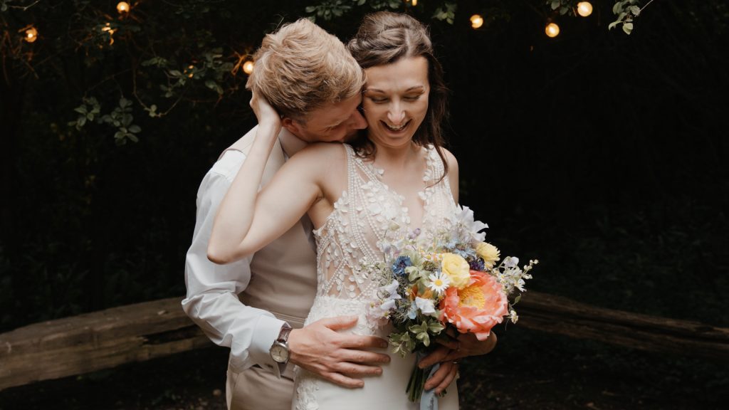 Groom romantically embraces bride from behind as she smiles holding a colourful wildflower bouquet during their Somerset wedding.