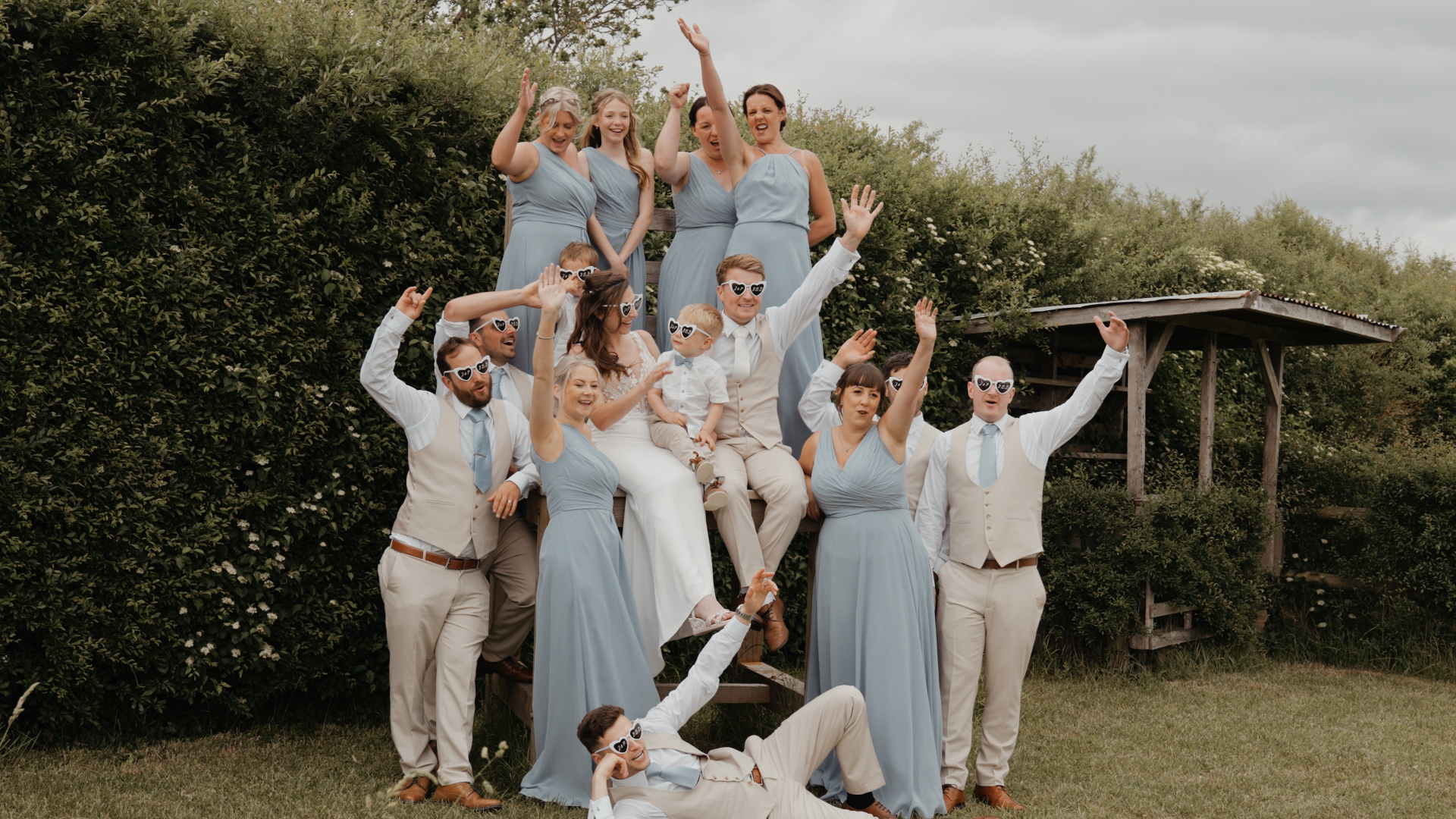 Newlyweds pose with bridesmaids, groomsmen, and their young son in a playful group photo at Old Oak Farm, wearing heart-shaped glasses.
