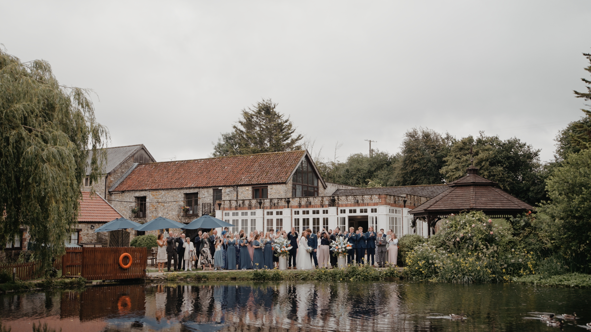 Guests celebrating outside Hornsbury Mill with the bride and groom by the water during their summer wedding.