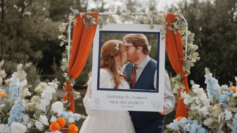 Newlyweds, Abi & Josh, sharing a kiss through a decorated photo frame surrounded by colourful summer flowers at Hornsbury Mill.