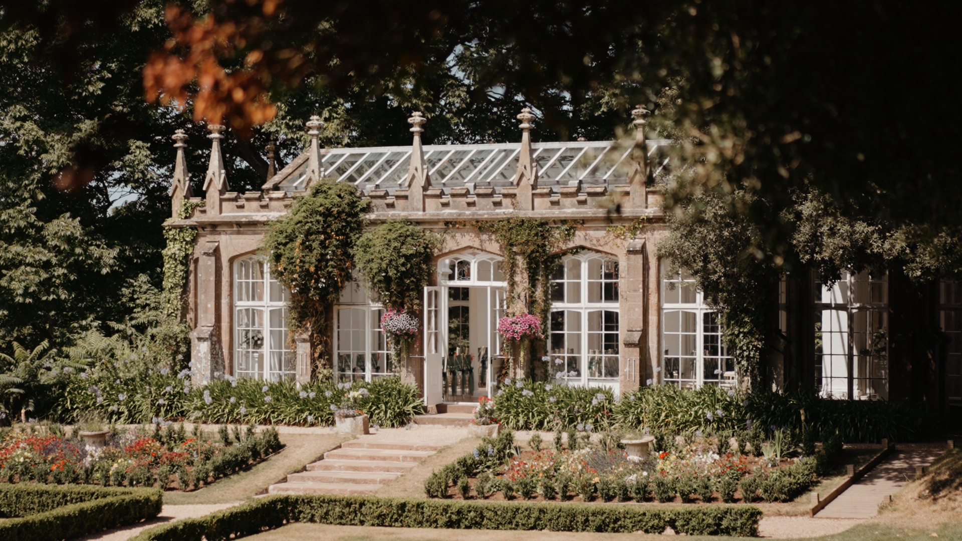 A scenic view of the glass orangery at St Audries Park, framed by gardens and ivy, ready for a summer wedding ceremony.