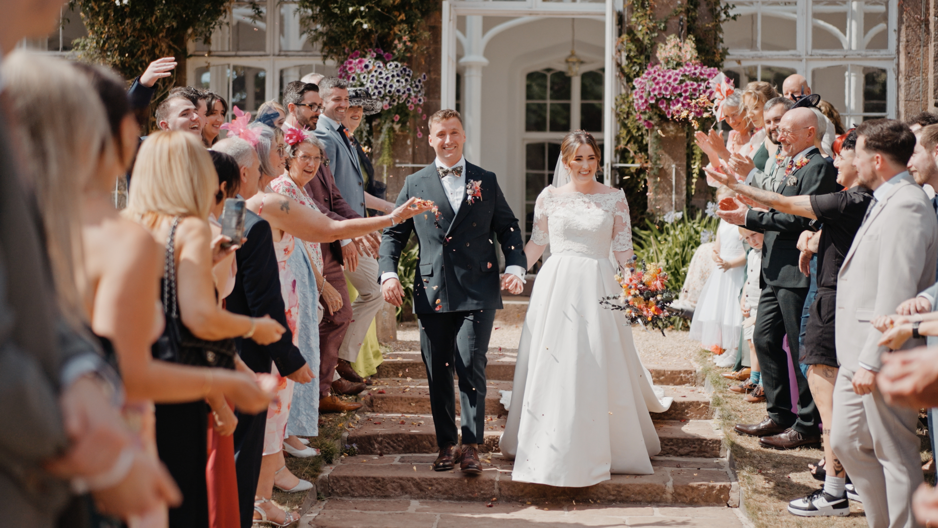The newlyweds walk through a colourful confetti toss line outside St Audries Park, surrounded by cheering guests.