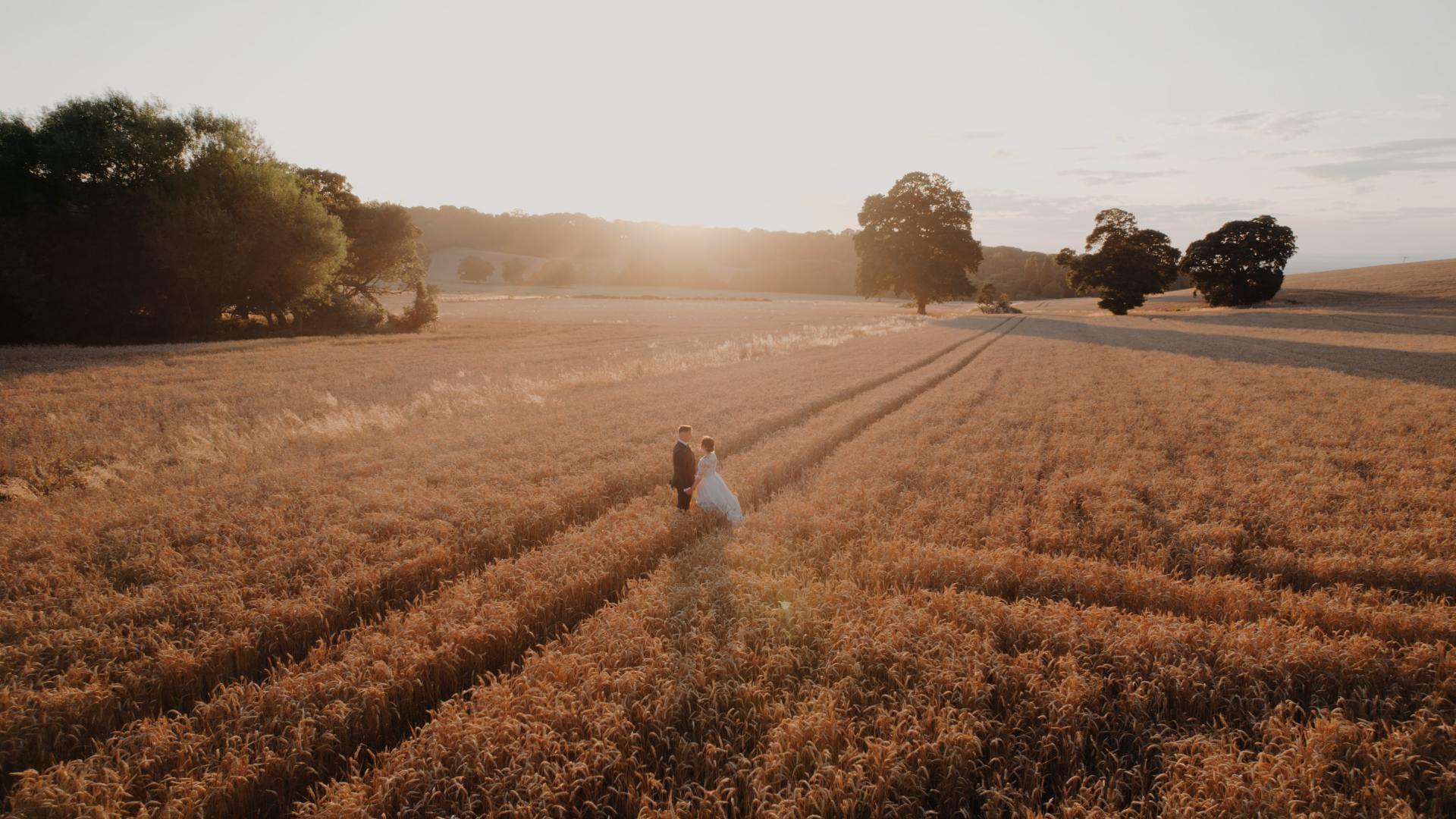 A golden hour couple portrait in a wheat field, the bride and groom walking hand in hand as the sun sets.