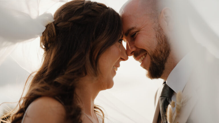 Close-up of the bride and groom under the veil, smiling softly with sunlight glowing through the fabric — a tender rustic wedding moment at Humber Barn.