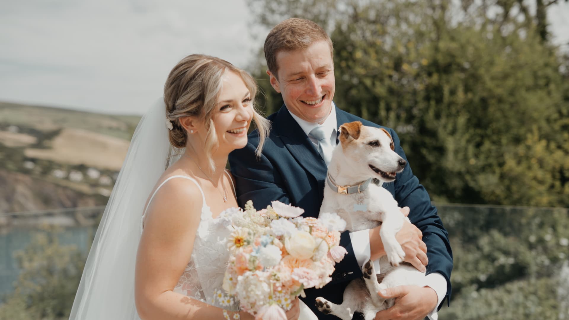 Bride and groom holding their dog while smiling on the terrace at Sandy Cove Hotel with coastal scenery behind them.