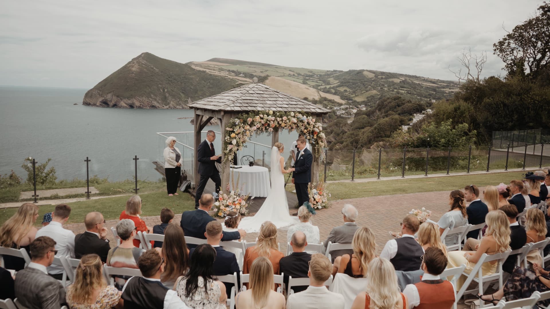 Bride and groom exchanging vows under floral arch at Sandy Cove Hotel in Devon, overlooking the sea.