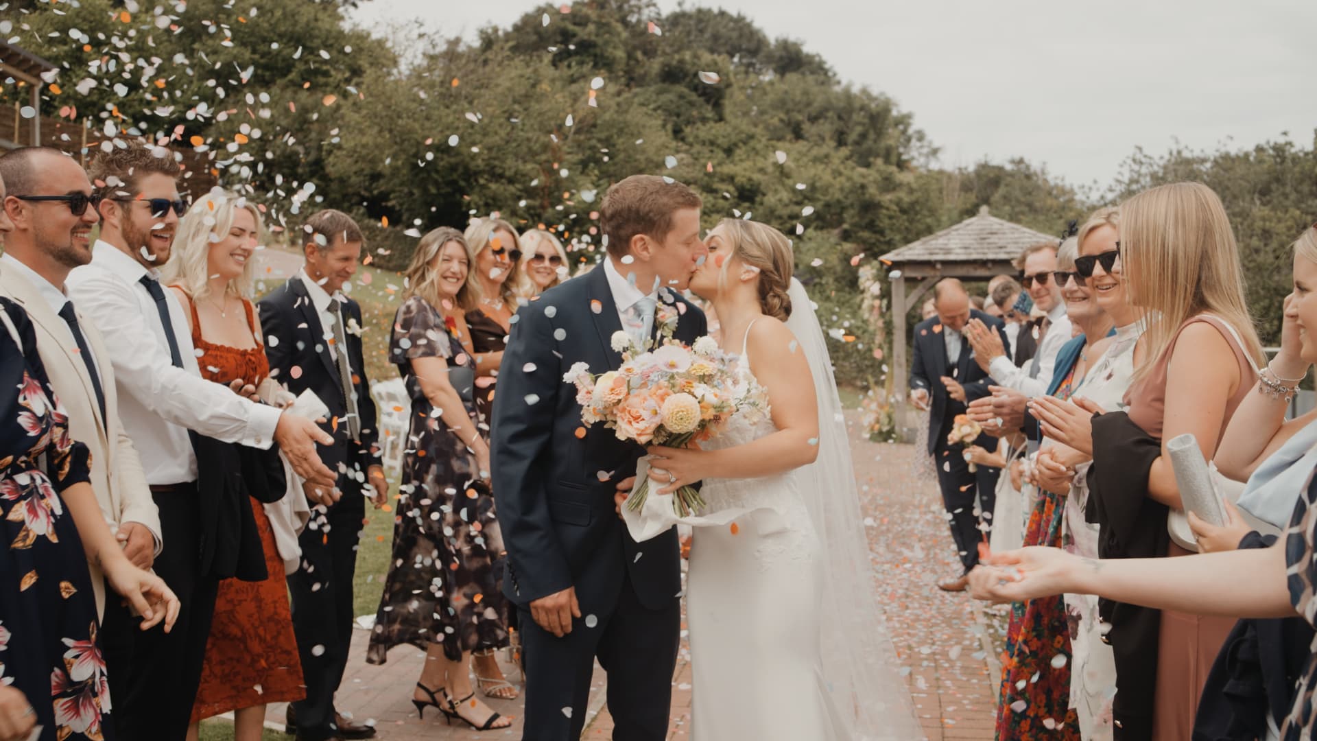 Newlyweds share a kiss as guests throw confetti outside Sandy Cove Hotel during their joyful summer wedding.