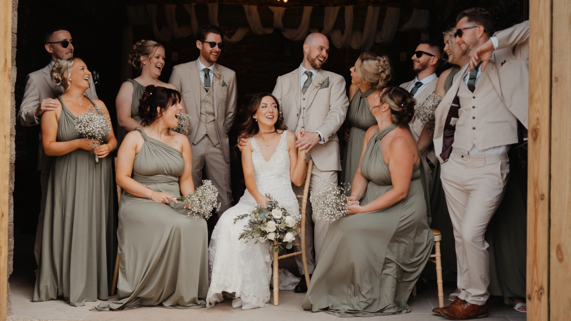 Bridal party and groomsmen laughing and celebrating with the bride and groom inside the barn, dressed in sage green bridesmaid dresses and beige suits — joyful rustic wedding vibes.