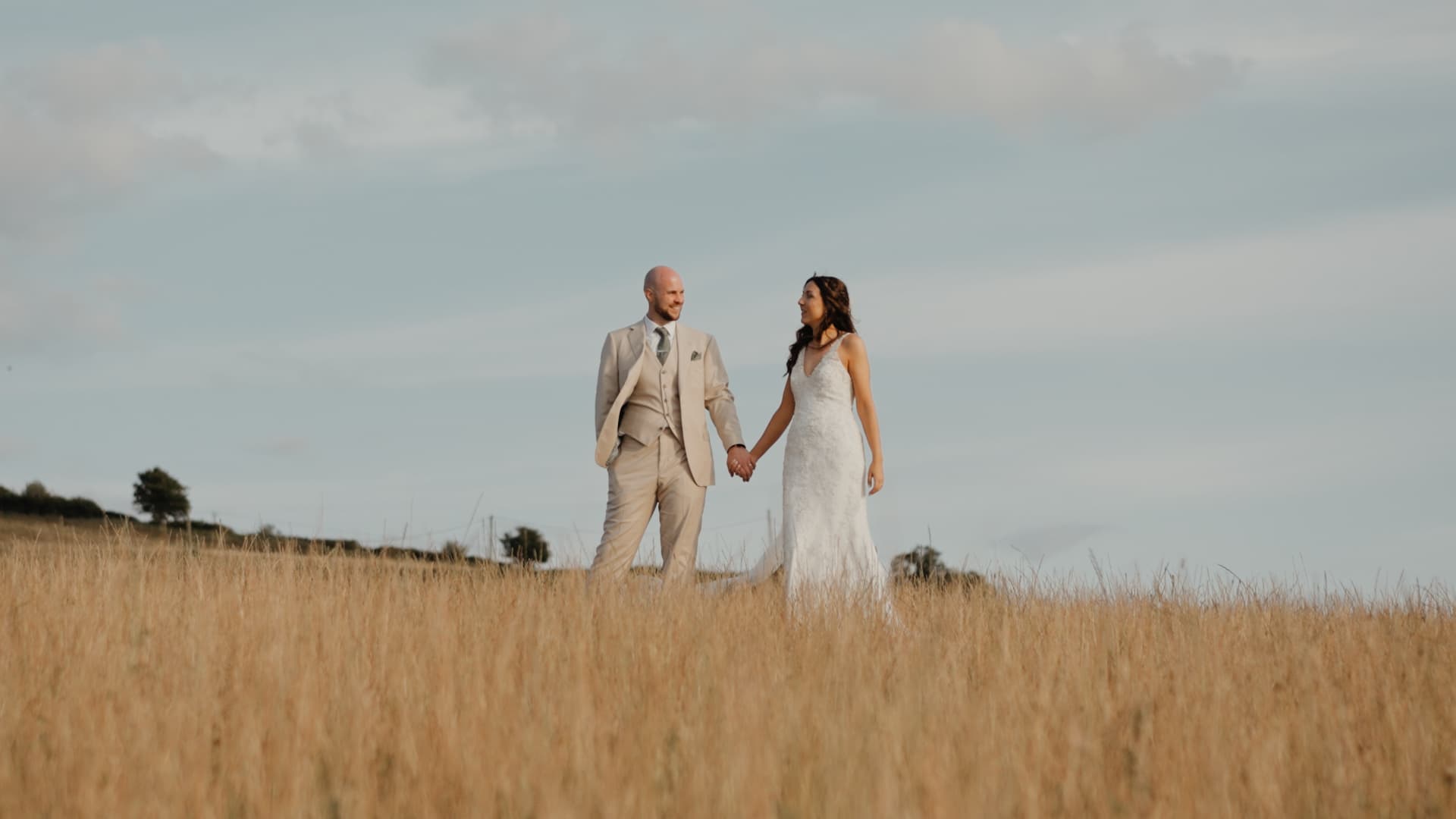 Bride and groom holding hands while walking through tall grass under soft natural light, capturing a romantic rustic wedding feel.