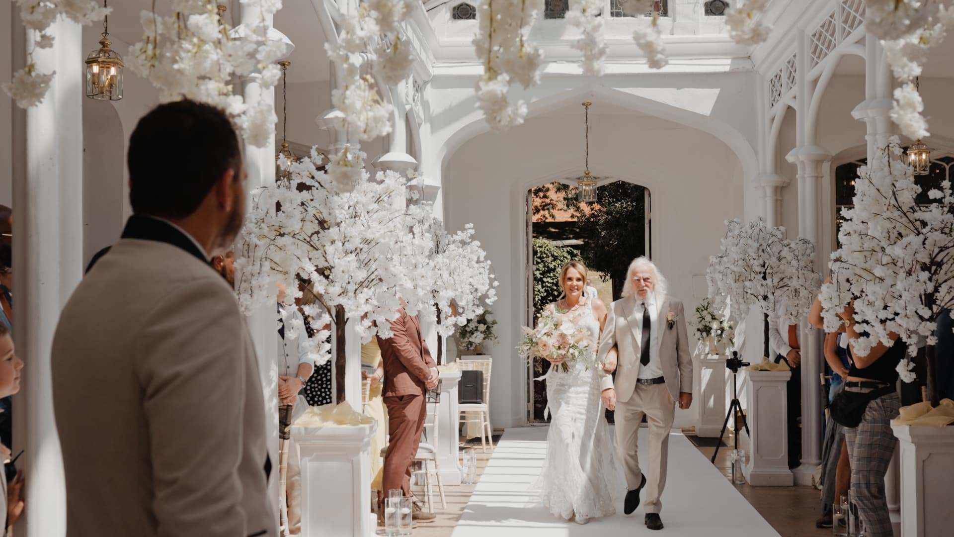 Bride walks down the aisle with her father in the bright Orangery at St Audries Park surrounded by guests and white florals.