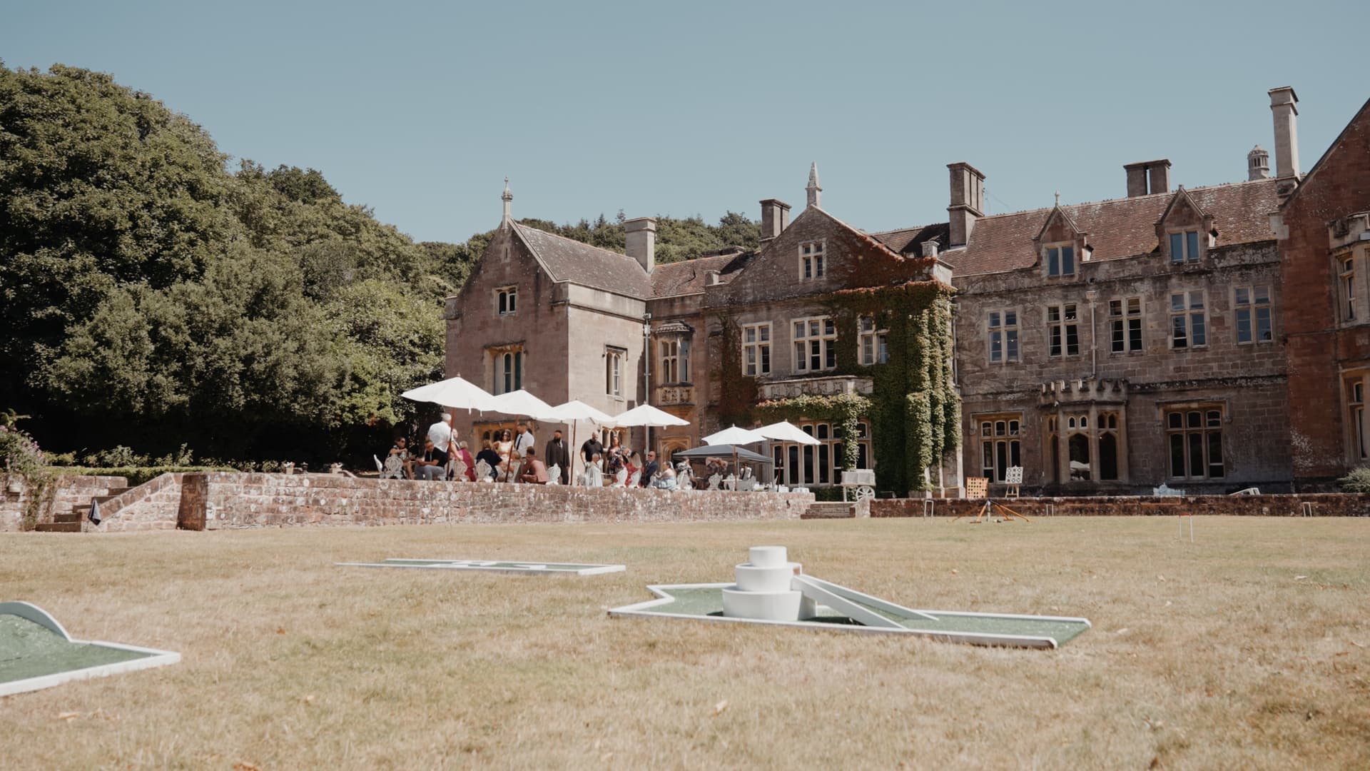 Guests enjoy drinks and conversation on the terrace at St Audries Park while lawn games like mini golf and ring toss sit ready on the sunny field.