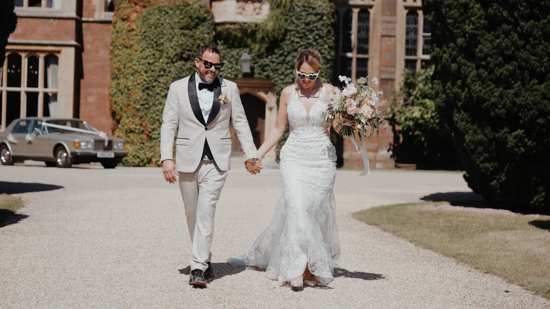 Bride and groom walk hand in hand outside St Audries Park, wearing chic sunglasses and modern wedding attire.