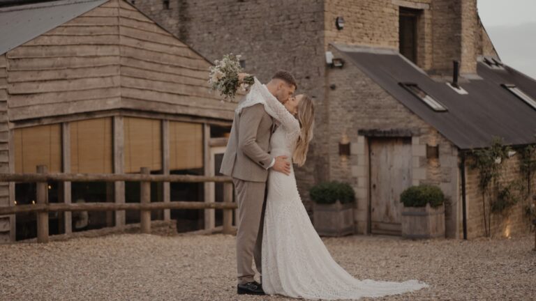 Romantic couple embrace outside Cripps Barn, surrounded by stone buildings that reflect Pendrell Hall’s timeless beauty.