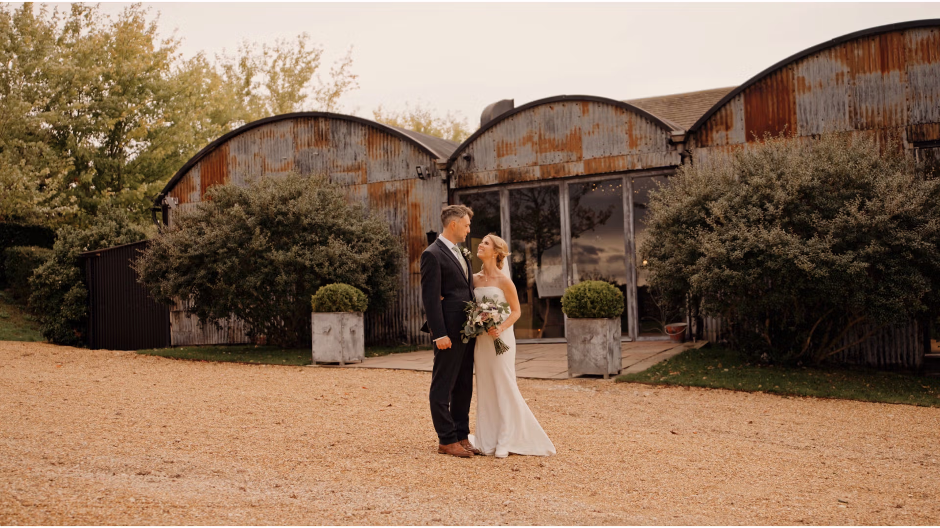 A bride and groom stand together outside a rustic building with corrugated metal walls. The groom is in a black suit, and the bride is in a white dress, holding a bouquet. The background features greenery and a gravel path.