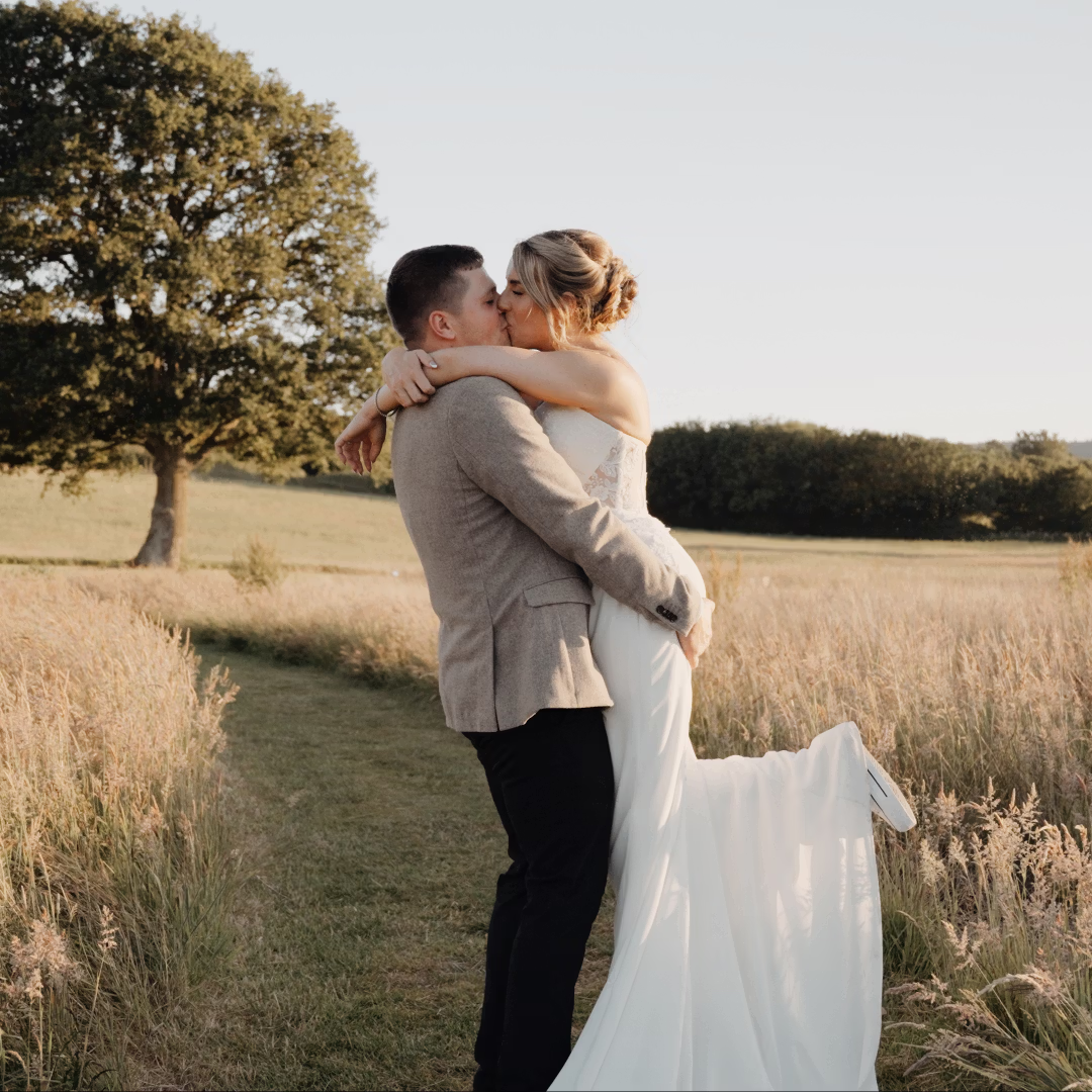 A couple embraces and kisses in a field. The woman, in a white dress, is lifted by the man, who wears a beige jacket and dark pants. They are surrounded by tall grass, with a large tree and distant hills in the background.