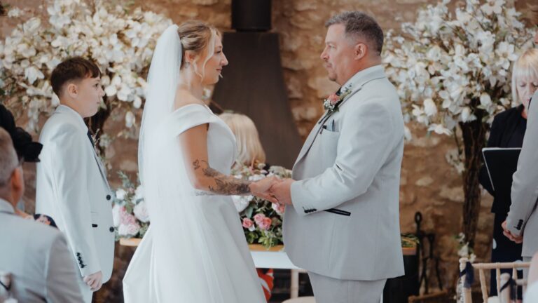 Bride and groom holding hands during their rustic barn ceremony at Quantock Lakes Wedding Venue, surrounded by soft florals and warm stone walls.