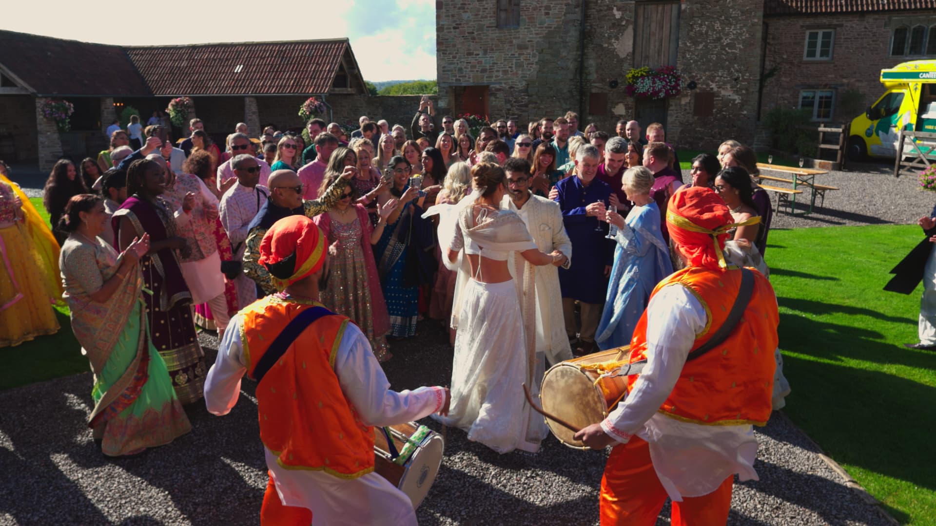 Wedding guests gathered around Dhol drummers during a lively rustic barn wedding, documented in a relaxed documentary style.