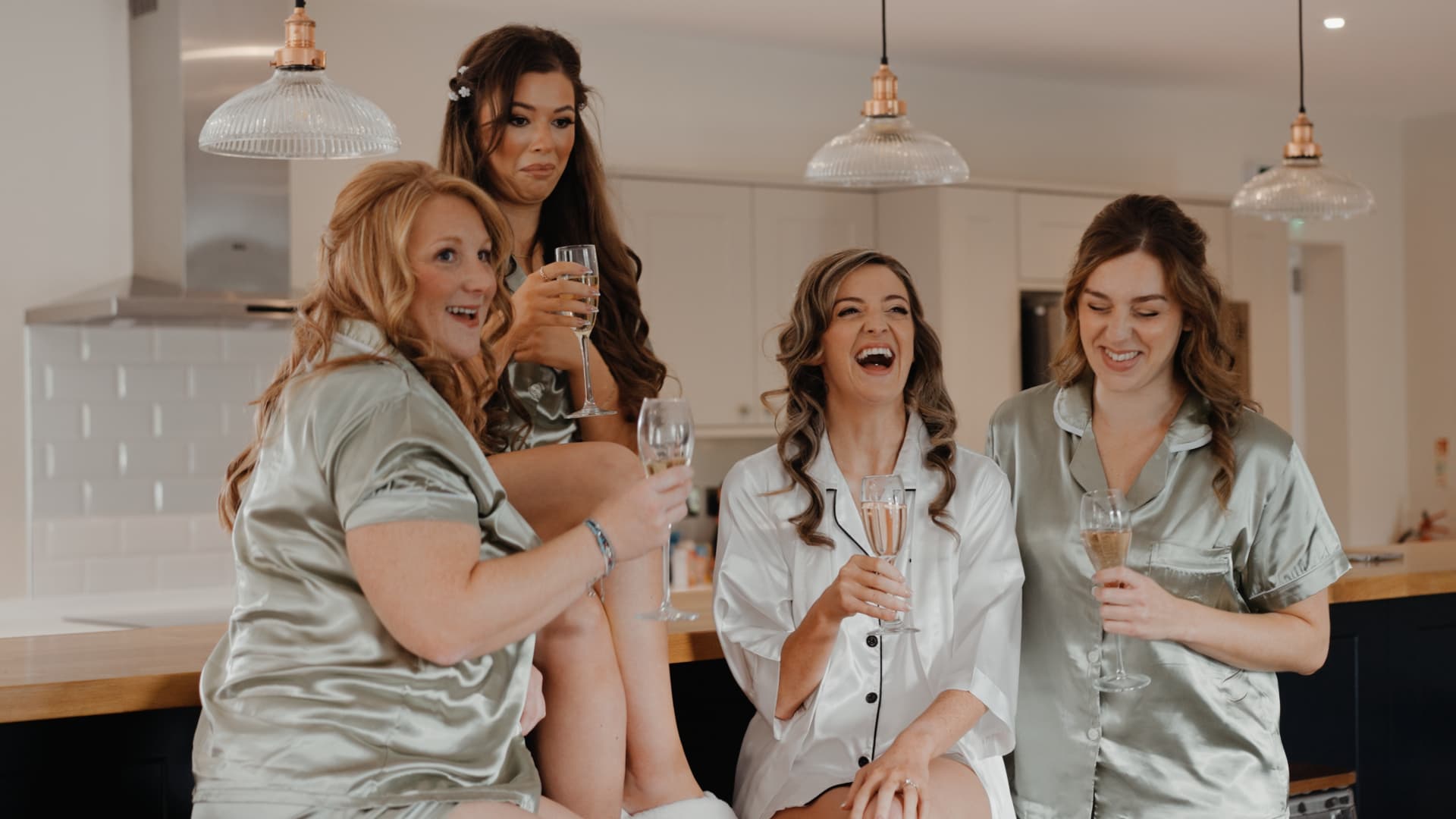 Bride and bridesmaids laughing together in matching satin pyjamas during relaxed morning preparations for their lakeside wedding film.
