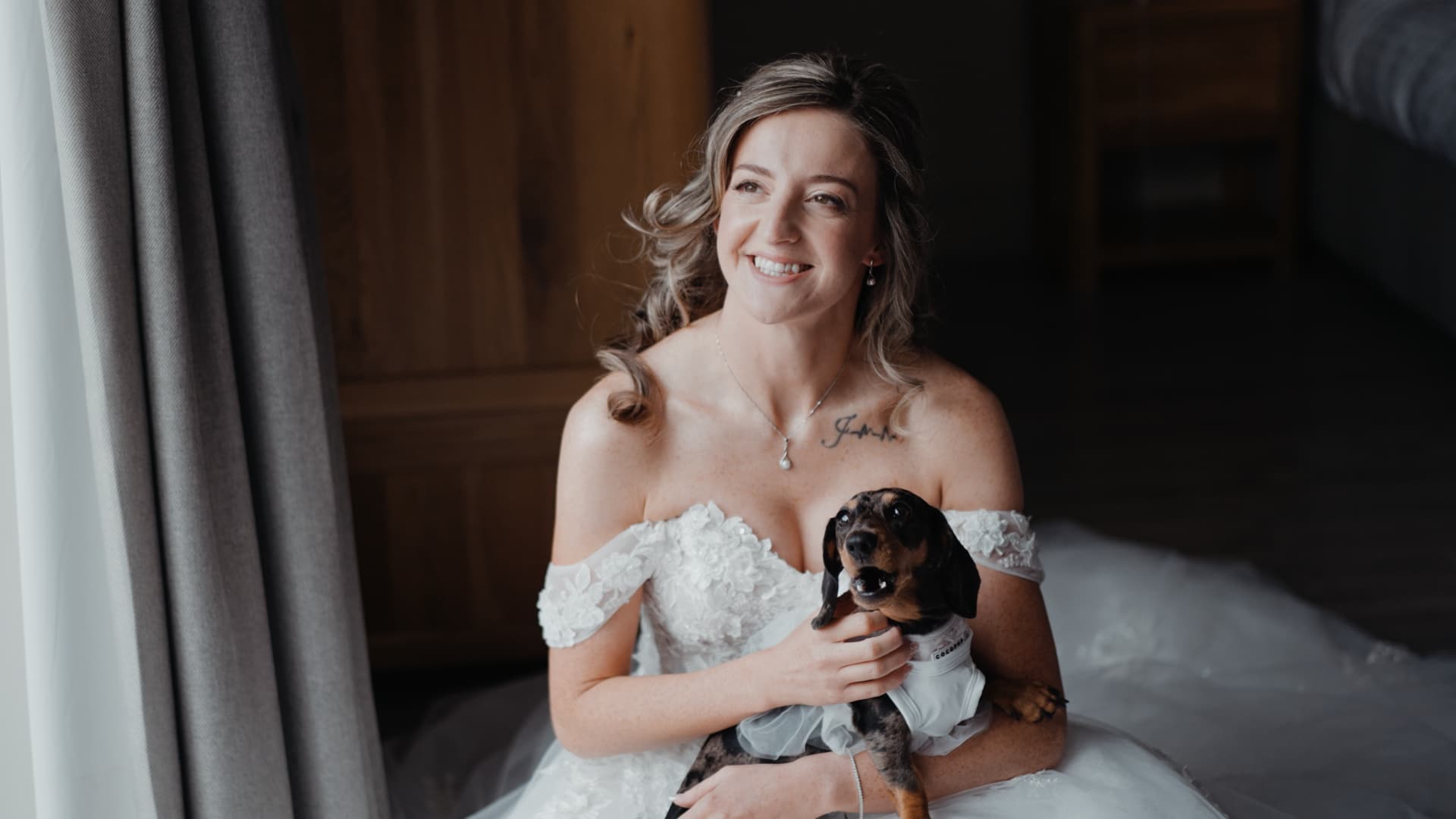 Bride smiling by the window while holding her dachshund during morning preparations, captured in a soft, natural style for their lakeside wedding film.