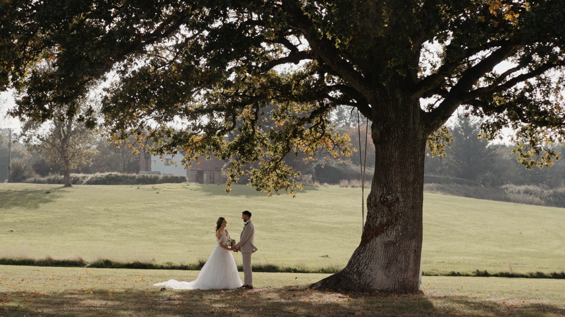Bride and groom standing together beneath a large oak tree during their rustic countryside wedding, framed by autumn light and open fields.