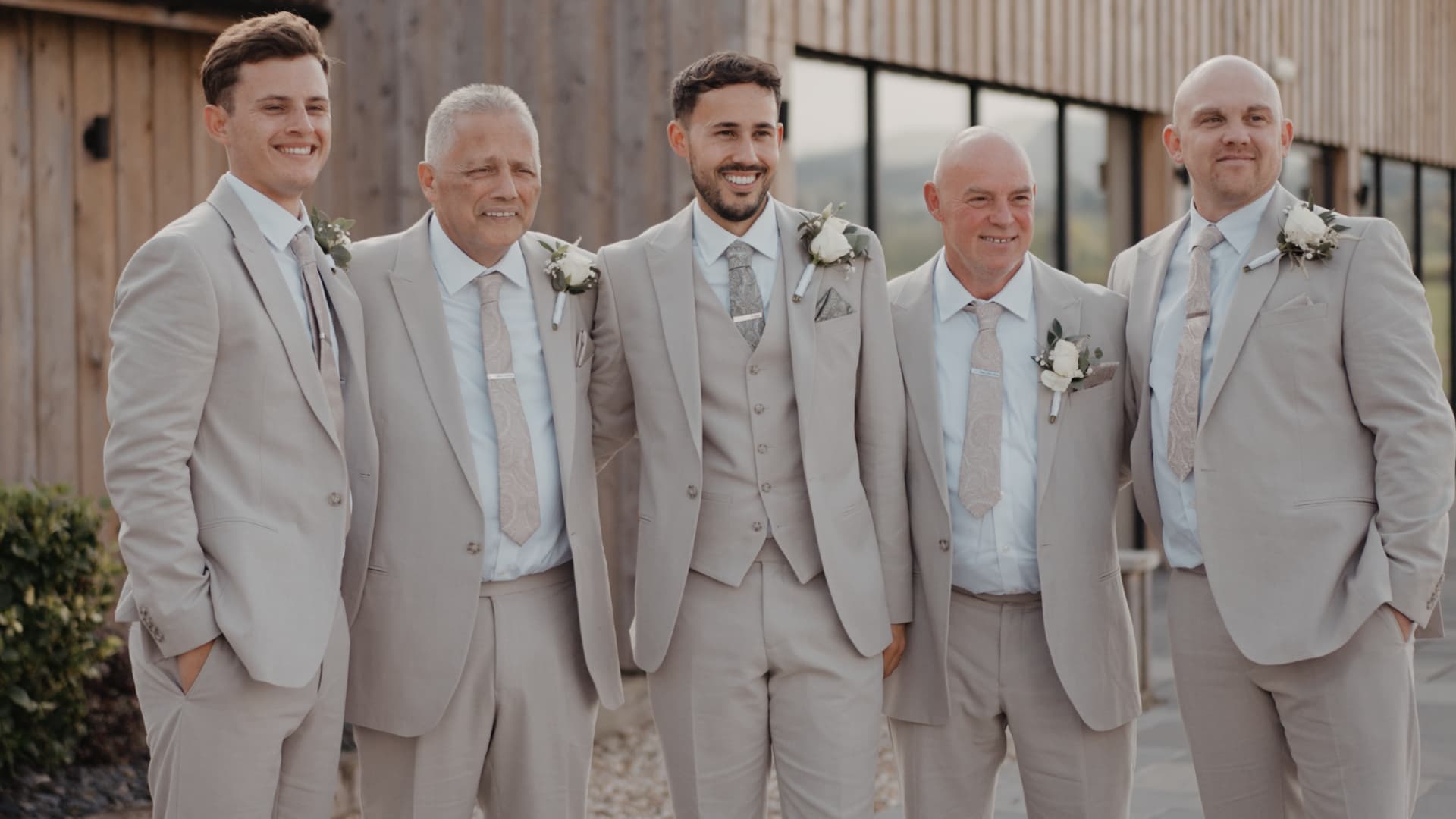 Groom standing with his groomsmen in matching neutral suits during their rustic countryside wedding, captured outside the barn.