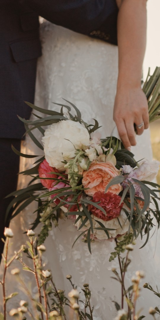 Bride and groom standing in a meadow at Kingscote Barn, Cotswolds, with bouquet detail captured in a luxury documentary wedding film by a Somerset wedding videographer