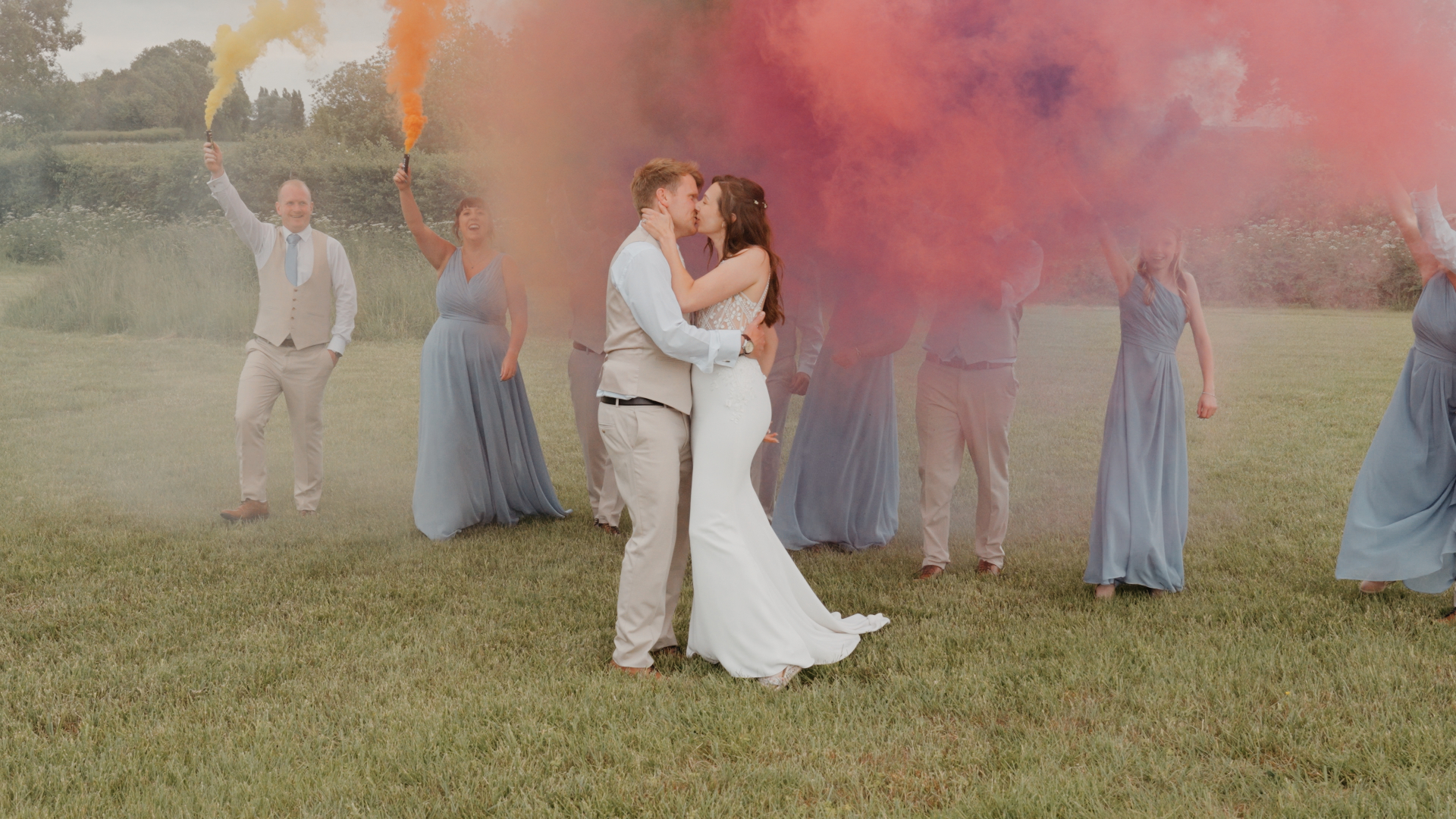 Bride and groom kiss surrounded by colourful smoke bombs in pink, orange, yellow, and purple at Old Oak Farm, with bridesmaids and groomsmen behind them.