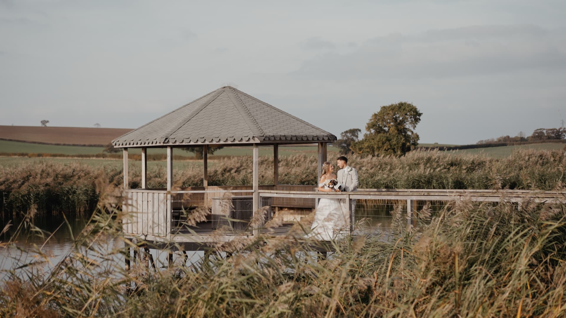 Bride and groom standing on a wooden lakeside gazebo at Quantock Lakes Wedding Venue, surrounded by tall reeds and open countryside views.