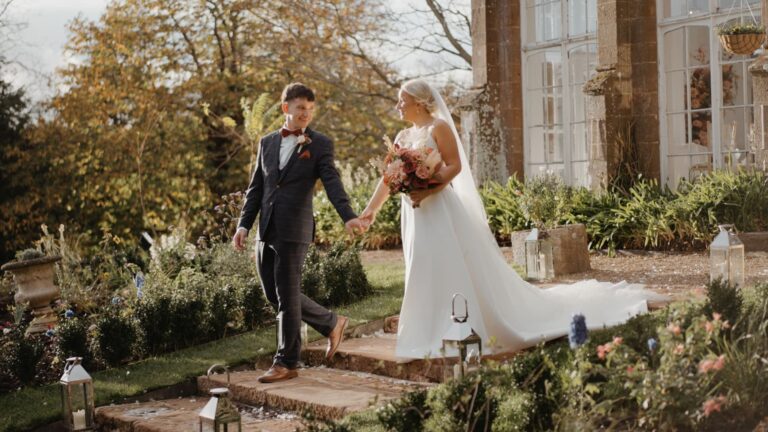 Newlyweds walk hand in hand through a country house garden, filmed by a Somerset wedding videographer