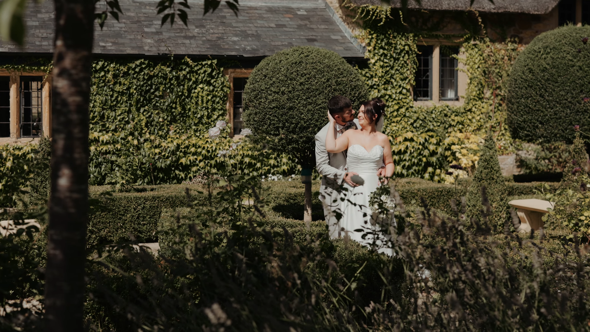 Aimee and Luke embrace in a formal Somerset garden beside an ivy-covered stone house on their wedding day